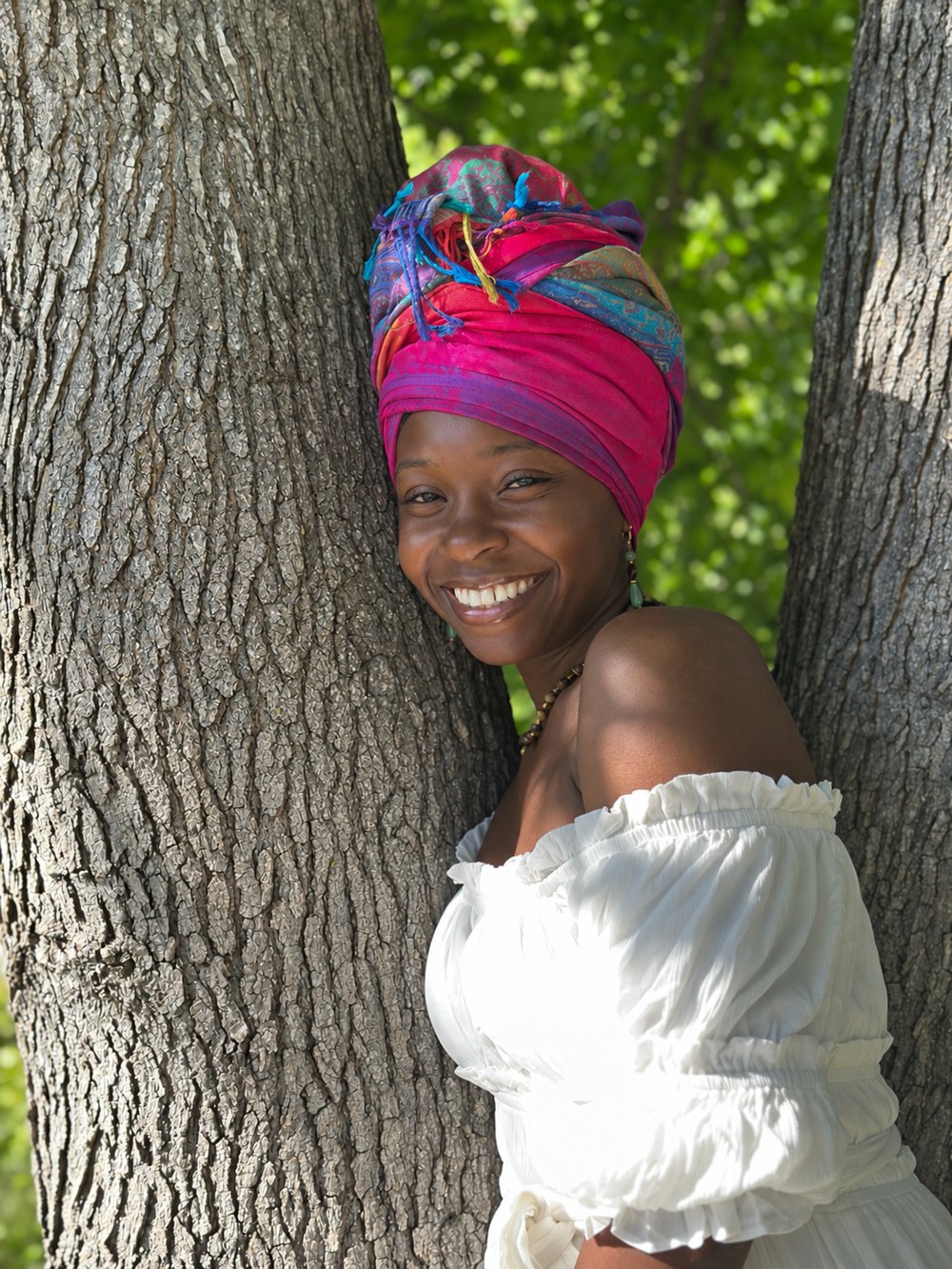 Kaitlen smiling in a pink head wrap and white dress, leaning against a tree